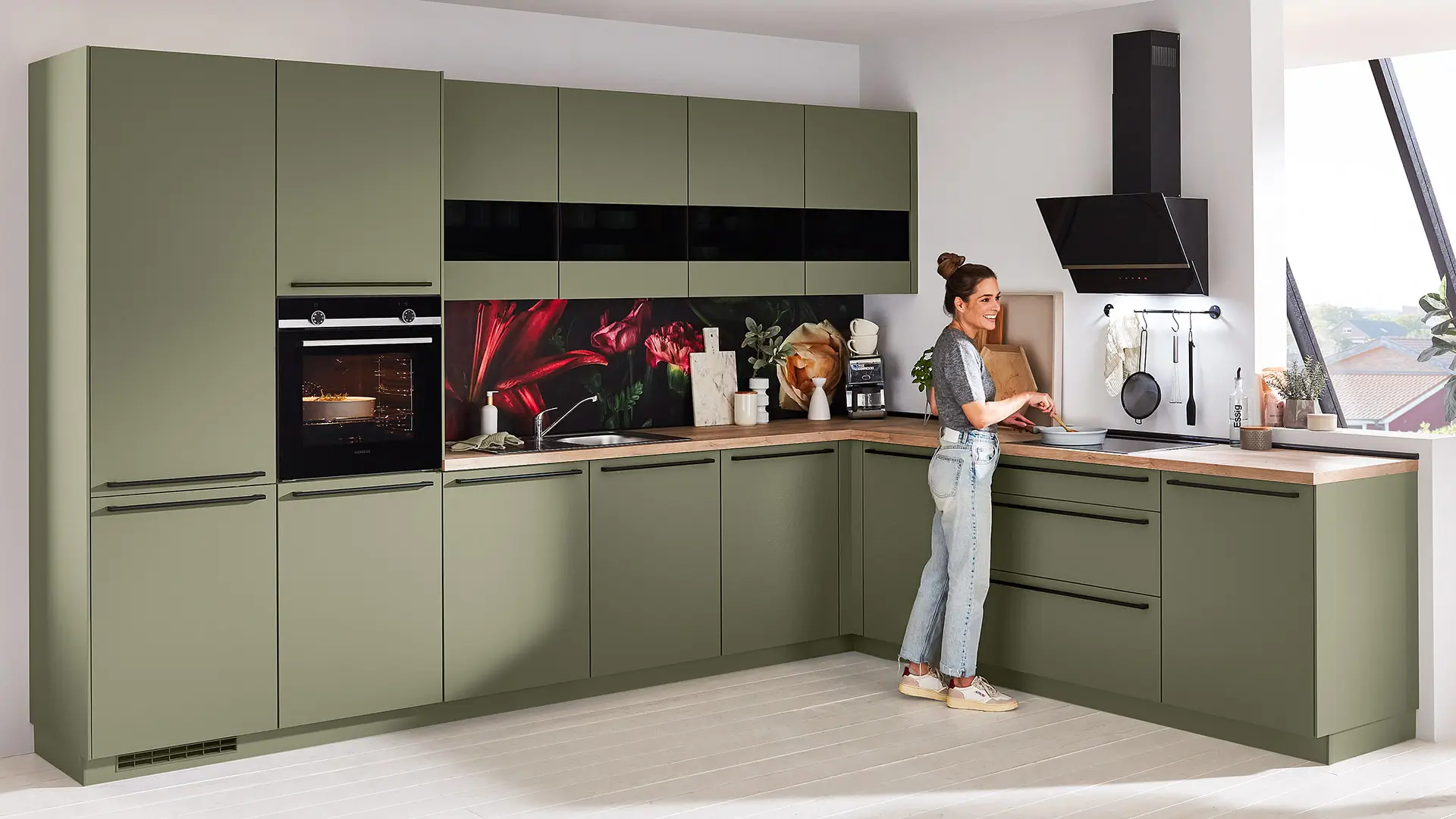 Frau kocht in moderner grüner Einbauküche mit Blumenrückwand und schwarzer Dunstabzugshaube.